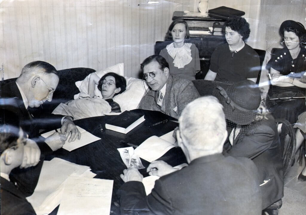 A black and white photograph showing a frail woman lying in bed, surrounded by lawyers and officials taking notes during what appears to be a legal hearing or deposition. Several women sit in the background, watching solemnly. The setting and context suggest this could be related to the 'Radium Girls' trials, where workers testified about radium poisoning.