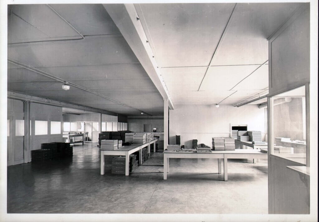 A black and white photograph of a large, empty factory or warehouse space with long tables stacked with organized trays and boxes. The room is well-lit, featuring a high ceiling with exposed lighting fixtures and a polished floor. The setting suggests an early-to-mid 20th-century industrial or assembly facility.