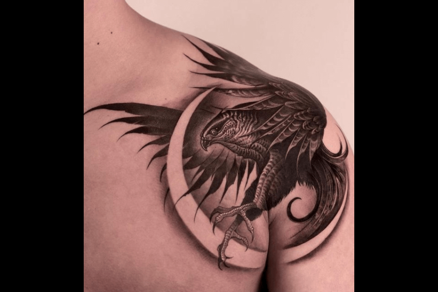 A highly detailed tattoo of a fierce bird, possibly a raven or hawk, in mid-flight with outstretched wings that extend across the shoulder and chest. The design incorporates intricate shading, emphasizing the bird's feathers, sharp beak, and talons, creating a sense of motion and power. A crescent moon frames the bird, adding an element of mysticism and elegance to the bold composition.