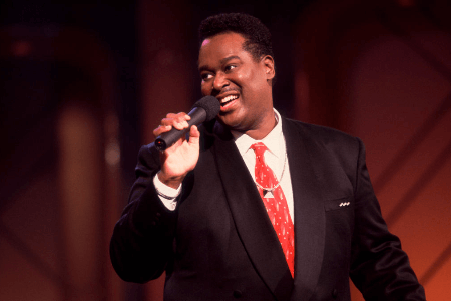 A male singer performs on stage, holding a microphone and smiling warmly. He is dressed in a black suit with a white shirt and a red tie adorned with subtle patterns. The stage lighting casts a warm glow, complementing his engaging and charismatic presence.