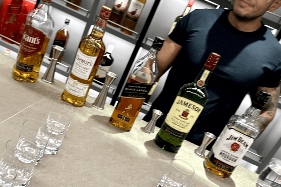 A bartender in a black t-shirt stands behind a counter displaying an assortment of whiskey bottles, including Jameson, Johnnie Walker Black Label, Dewar’s, Jim Beam, and Grant’s. Several shot glasses and metal jiggers are arranged on the countertop, indicating a whiskey tasting or bar service. The background features shelves stocked with additional liquor bottles.