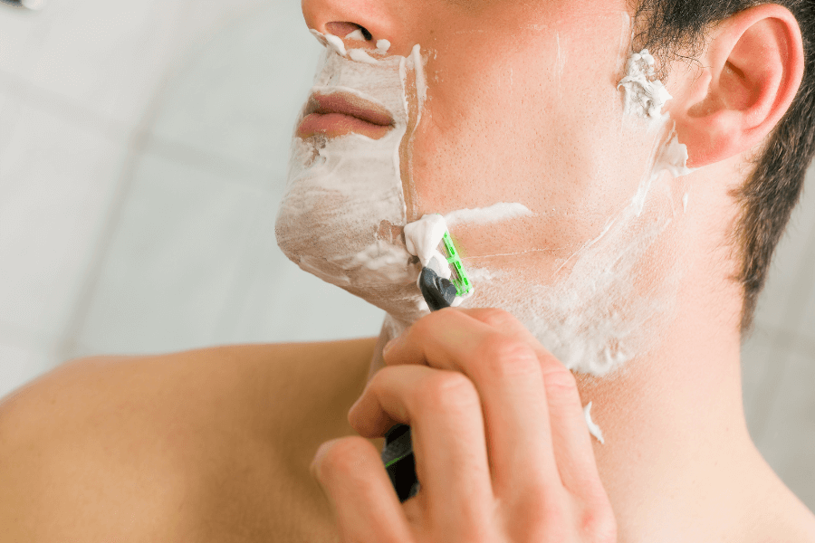 A close-up of a man shaving his face with a razor, covered in thick white shaving cream. The razor glides along his cheek, revealing smooth skin beneath the foam.