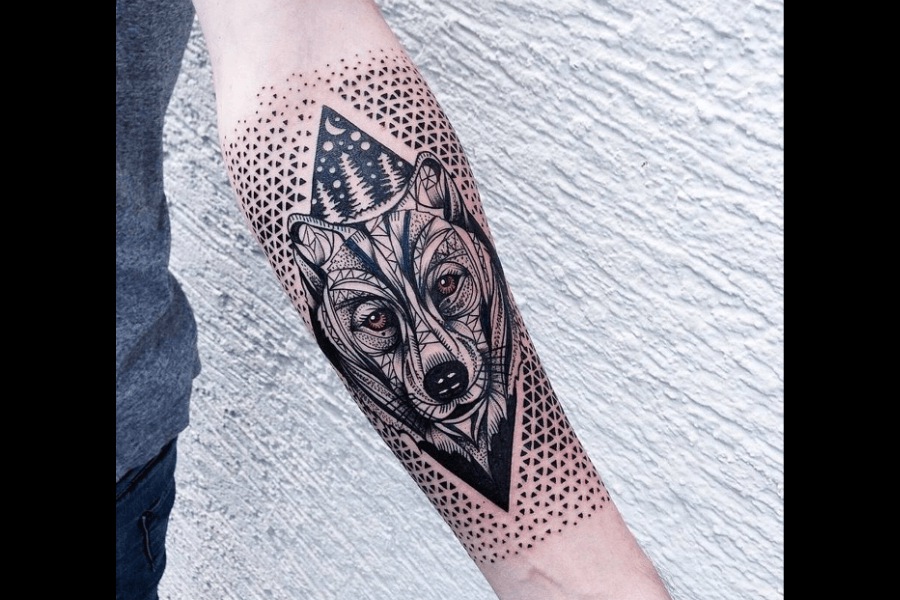 A geometric tattoo featuring a wolf's face surrounded by intricate line work and patterns. The background includes a triangle frame and dotted designs, with trees subtly incorporated into the upper triangle.