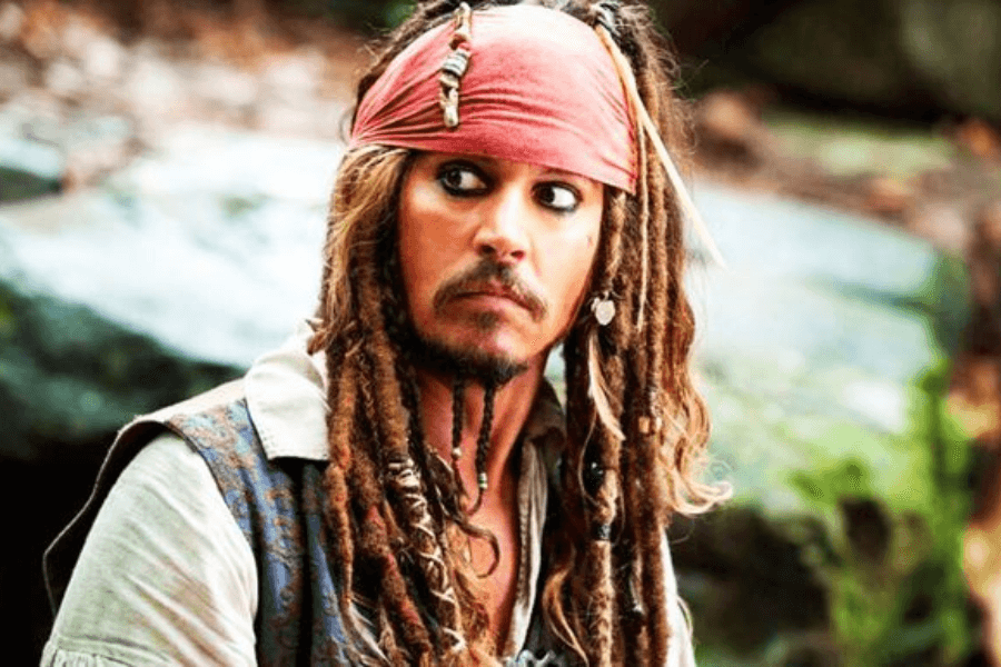 A pirate with long, beaded dreadlocks and dark kohl-lined eyes wears a red bandana and a weathered vest over a white shirt. He has an intense and slightly surprised expression as he looks off to the side, standing in a lush, outdoor setting with blurred greenery and rocks in the background. His rugged attire and accessories give him a swashbuckling, adventurous appearance.