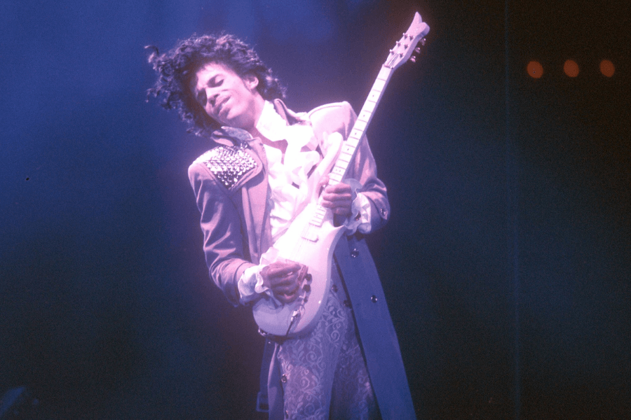 A male guitarist performs passionately on stage, dressed in a ruffled white shirt, a purple embellished jacket, and matching patterned pants. His curly hair bounces as he leans into his guitar, eyes closed, fully immersed in the music. The stage lighting casts a dramatic blue and purple glow, enhancing the electrifying atmosphere.