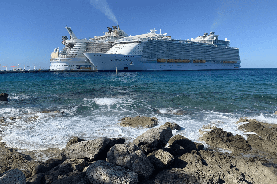 Two massive Royal Caribbean cruise ships, including Harmony of the Seas, docked at a port with clear blue ocean water and rocky shoreline in the foreground. Smoke rises from the ship's funnels as the vessels tower over the pier. The bright blue sky and gentle waves create a picturesque seaside scene.