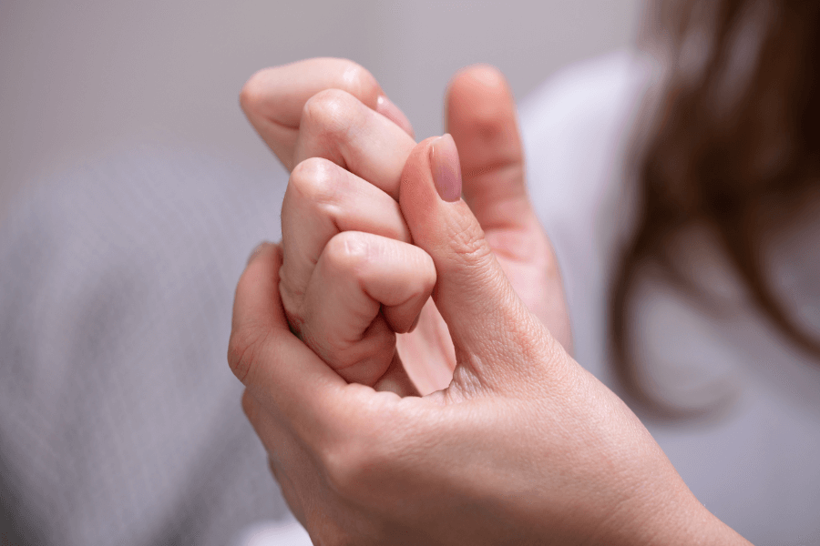 A close-up of a person’s hands as they press their fingers together, seemingly cracking their knuckles. The background is blurred, with a hint of long hair and a white shirt visible.