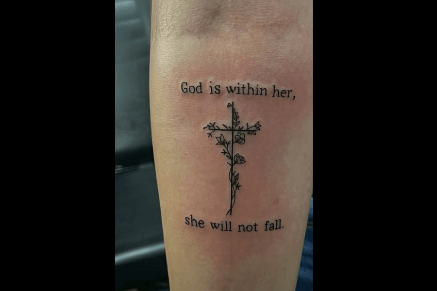 A simple tattoo of a cross made of floral stems and buds. The text reads, “God is within her, she will not fall,” placed above and below the cross.