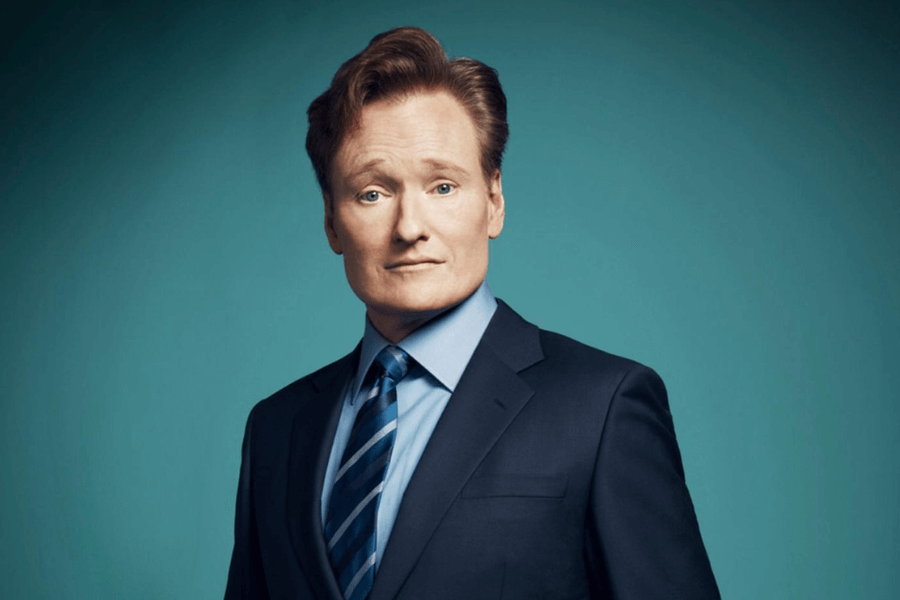 A man with neatly styled reddish-brown hair stands against a teal background, wearing a dark navy suit, a light blue dress shirt, and a striped blue tie. He has a slightly raised eyebrow and a subtle smirk, giving him a charismatic yet composed appearance. The lighting is soft and even, emphasizing his features.