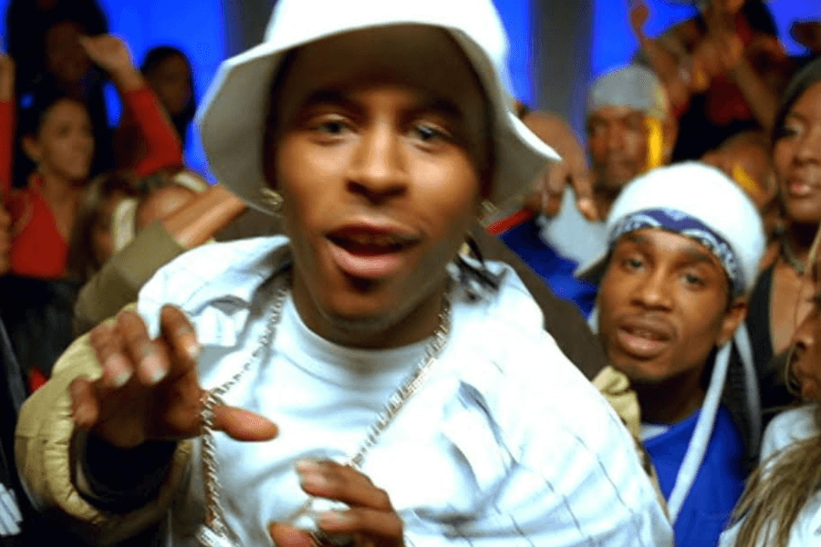 A man wearing a white bucket hat, a white t-shirt, and a gold chain leans toward the camera with an energetic expression in a lively party scene. Behind him, another man in a white beanie and blue bandana smiles, while a crowd of people dances and raises their hands in the background. The image has a vibrant, early 2000s hip-hop music video aesthetic.