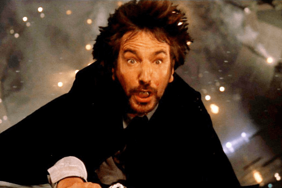 A man in a black suit with disheveled hair and a shocked expression is falling through the air, his mouth open in fear. The background is blurred with sparks and debris, suggesting an explosion or dramatic action scene. His outstretched arm and the intense lighting add to the sense of motion and suspense.