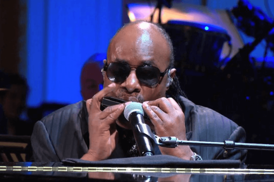 A male musician wearing dark sunglasses and a black suit plays the harmonica while seated at a piano. He leans into the microphone, deeply engrossed in the performance, as stage lighting casts a cool blue glow around him. The blurred background reveals a live band setup, enhancing the concert atmosphere.