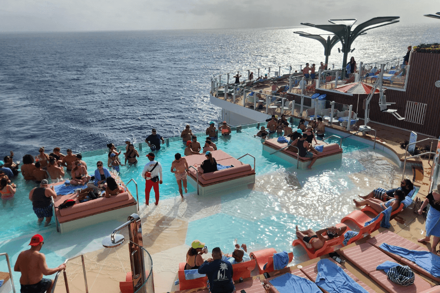 A luxurious infinity pool on a cruise ship filled with passengers relaxing in the water and on submerged lounge beds. Sunbathers recline on cushioned chairs while others socialize in the pool, with the open ocean stretching to the horizon in the background. The scene is lively and vibrant, with a mix of shaded and sunlit areas.