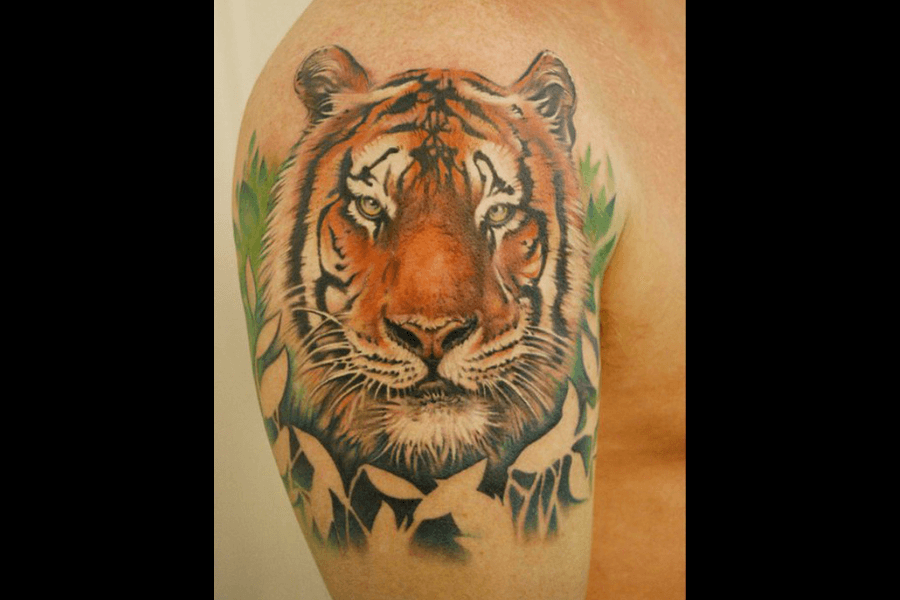A detailed tattoo of a tiger’s face with vibrant orange and black stripes. The background features green foliage and abstract shading, creating a jungle-like vibe.