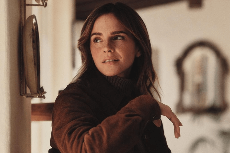 A young woman with shoulder-length brown hair gazes off to the side with a soft, thoughtful expression. She wears a brown suede jacket over a dark turtleneck, and the warm, dim lighting gives the setting a cozy, vintage feel. The blurred background features a mirror and rustic interior elements, enhancing the intimate and elegant atmosphere.