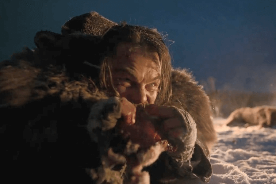 A rugged, fur-clad man with a weathered face and intense eyes bites into raw meat while lying in the snow. His expression shows desperation and determination, with a fire glowing in the distance, casting light on the icy wilderness. The harsh, cold environment and his survivalist appearance add to the raw, primal intensity of the moment.