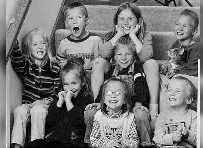 A black-and-white photo of eight young children sitting on a staircase, all smiling, laughing, or making playful faces. One child holds a small kitten, while others display various expressions of joy and excitement.