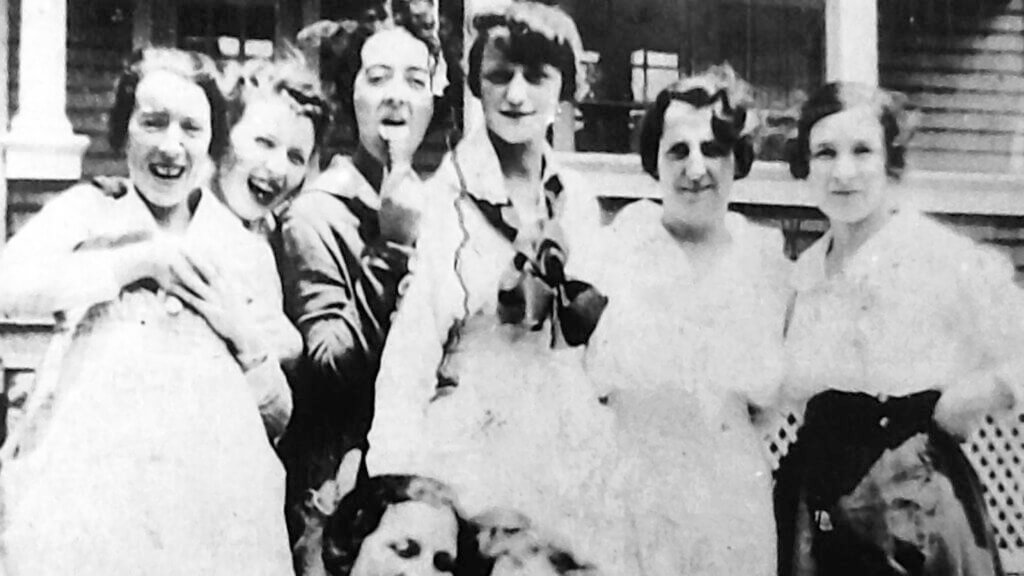 A black and white photograph of a group of six young women from the early 20th century, smiling and posing playfully. Some appear to be licking or pointing at paintbrushes, possibly referencing the 'Radium Girls,' factory workers who unknowingly ingested radium while painting luminous watch dials.