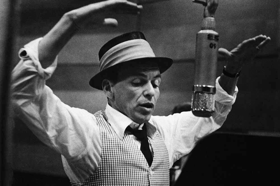 A black and white photograph of a male singer in a recording studio, wearing a fedora and a checkered vest over a white dress shirt. He raises both hands expressively while singing into a vintage microphone, capturing a moment of deep emotion and musical interpretation. The blurred studio background adds to the timeless and intimate atmosphere.