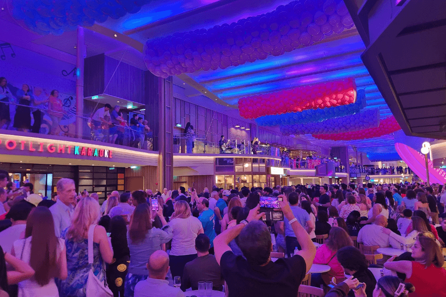 A vibrant indoor event on a cruise ship with a large crowd gathered under colorful balloon decorations and dynamic blue and purple lighting. Passengers watch a performance on an elevated stage while some record the moment on their phones. The venue features multiple levels, with a "Spotlight Karaoke" sign visible on the left, suggesting a lively entertainment area.