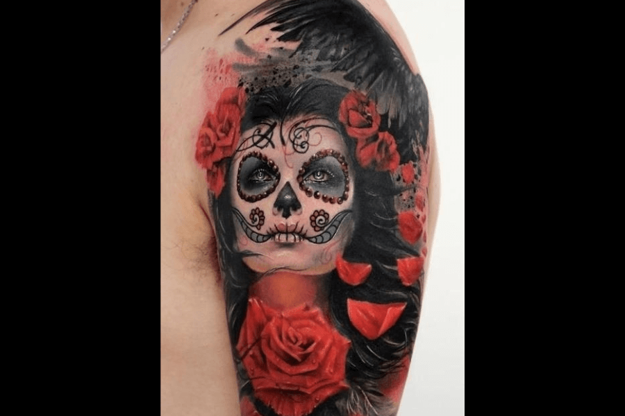A vibrant tattoo featuring a Day of the Dead-inspired sugar skull woman. The design includes intricate face details, bright red roses, and dark shading, with black feathers adding depth.