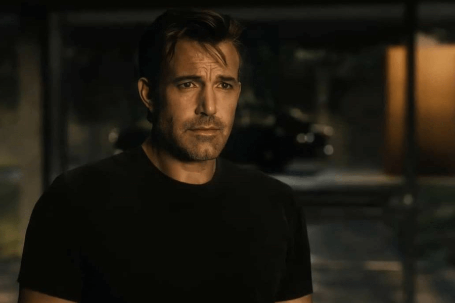 A man with short, slightly tousled brown hair and light stubble stands in a dimly lit setting, wearing a plain black T-shirt. His expression is serious and contemplative, with furrowed brows and a distant gaze. The background is blurred, featuring warm lighting and a faint outline of a car and modern architecture.