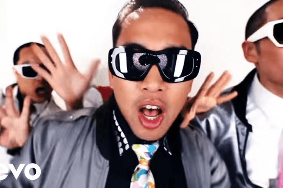 A man wearing oversized futuristic sunglasses, a silver jacket, and a colorful tie sings energetically into the camera with his mouth open. Behind him, two backup performers in matching silver outfits and white sunglasses strike dynamic hand gestures. The bright, high-contrast background and bold fashion choices give the image a playful, high-energy music video aesthetic.