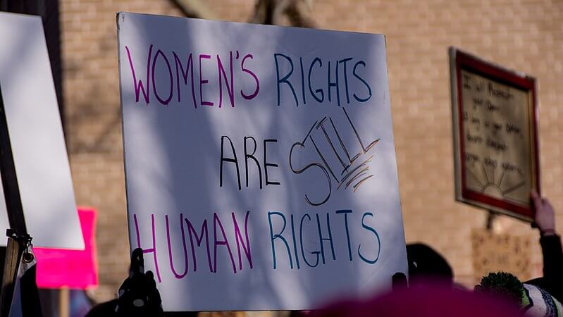 A close-up of a protest sign reading 'WOMEN'S RIGHTS ARE STILL HUMAN RIGHTS' in colorful, hand-drawn letters. The sign is held up during a demonstration, with other signs and protesters visible in the blurred background. The setting appears to be outdoors near a brick building.