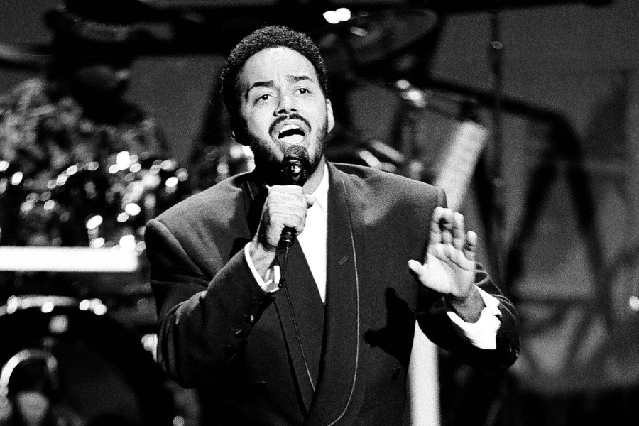 A black and white photograph of a male singer passionately performing on stage, holding a microphone in one hand while gesturing with the other. He wears a dark suit with a white shirt, his face expressive as he sings. The blurred background features a live band, adding to the dynamic concert atmosphere.