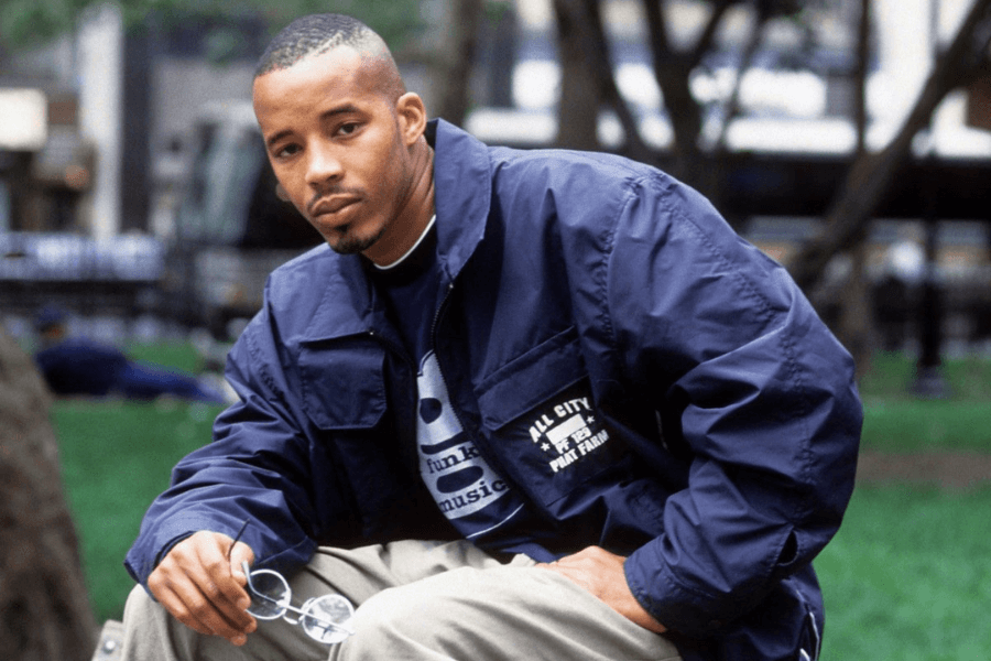 A man sitting in an urban park wearing a navy blue Phat Farm "All City" jacket over a graphic T-shirt, paired with khaki pants. He holds a pair of glasses in one hand and gazes at the camera with a confident yet relaxed expression. The outfit reflects the clean, streetwear-inspired style of the late '90s and early 2000s.