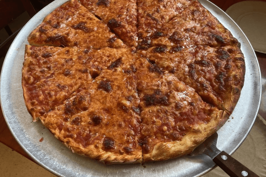  A freshly baked cheese pizza with a golden, bubbly, and slightly charred crust sits on a round metal tray. The pizza is cut into eight equal slices, with a generous layer of melted cheese over a tomato sauce base. A metal spatula is partially visible under one of the slices, ready for serving.