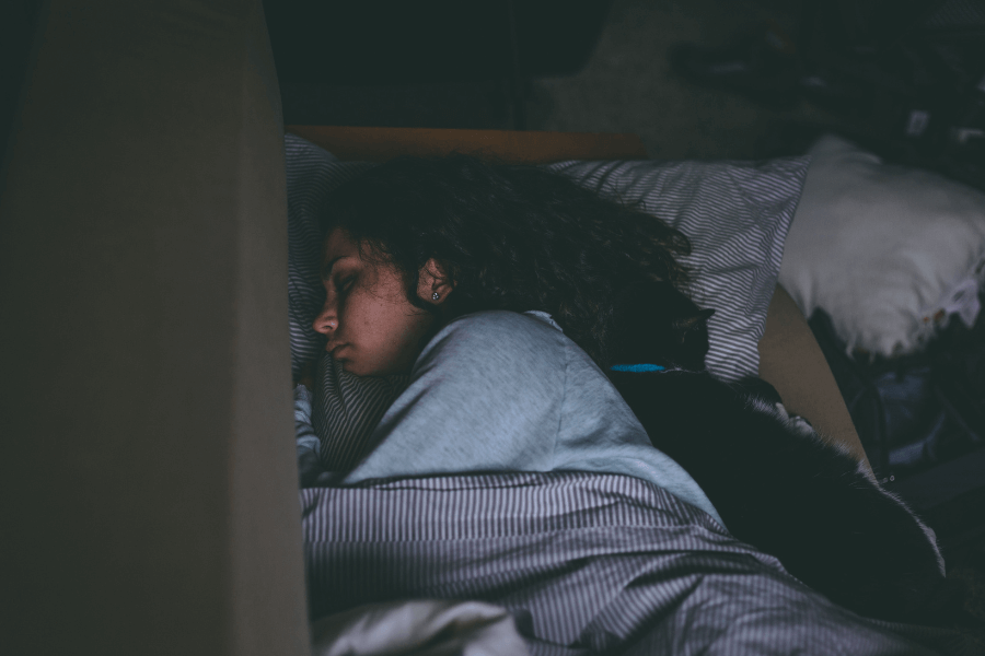 A woman with curly hair sleeps on her side in a dimly lit room, wrapped in a blanket, with a black and white cat snuggled against her back. The cozy setting is enhanced by soft lighting and a slightly messy background.