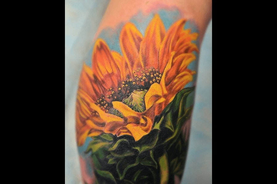 A vibrant tattoo of a sunflower in full bloom. The design features rich yellow petals, a detailed green stem, and a bright blue sky background.