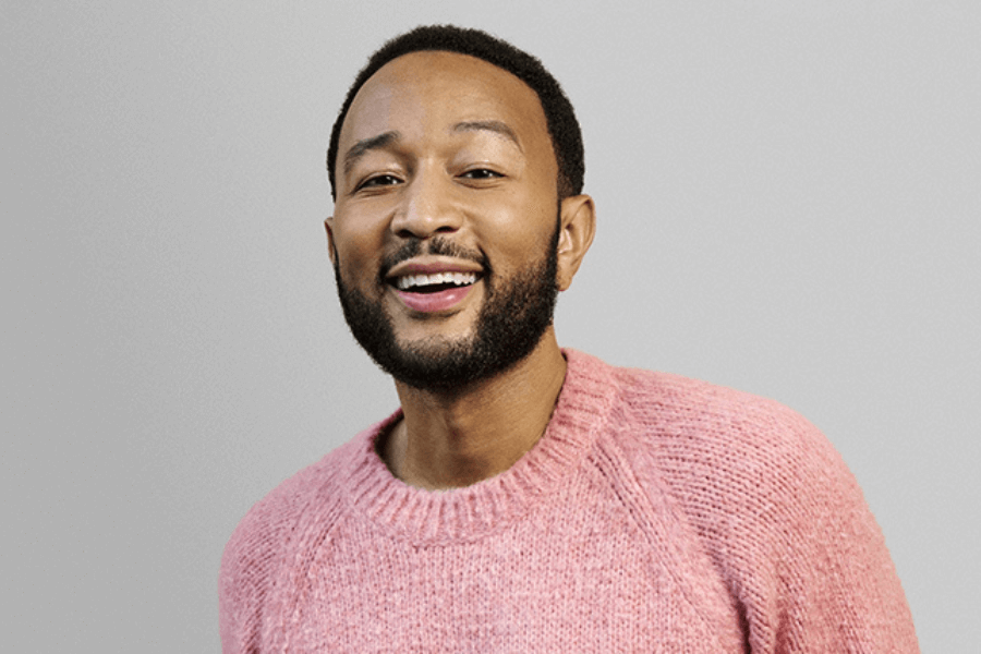 A smiling man with short black hair and a well-groomed beard wears a cozy pink knitted sweater against a plain light gray background. His warm expression and slightly tilted posture give off a friendly and approachable vibe. The soft lighting highlights his features and the texture of the sweater.