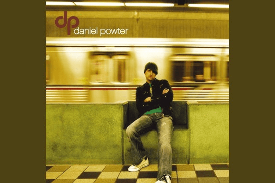 A man wearing a beanie, black jacket, and jeans sits on a subway station bench with his arms crossed, gazing toward the camera. Behind him, a train speeds past, creating a motion blur effect that contrasts with his stillness. The name "Daniel Powter" and his initials "dp" are displayed in the upper left corner, indicating this is an album cover.