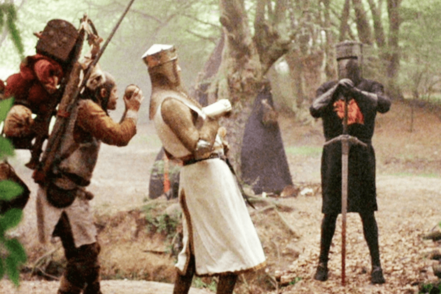 A medieval knight in black armor, known as the Black Knight, stands in a wooded area gripping a large sword in front of him. Facing him is a king dressed in white and gold armor, leaning back slightly, while a servant behind him holds two coconut halves. The scene is set in a misty forest, evoking a comedic yet dramatic medieval duel.