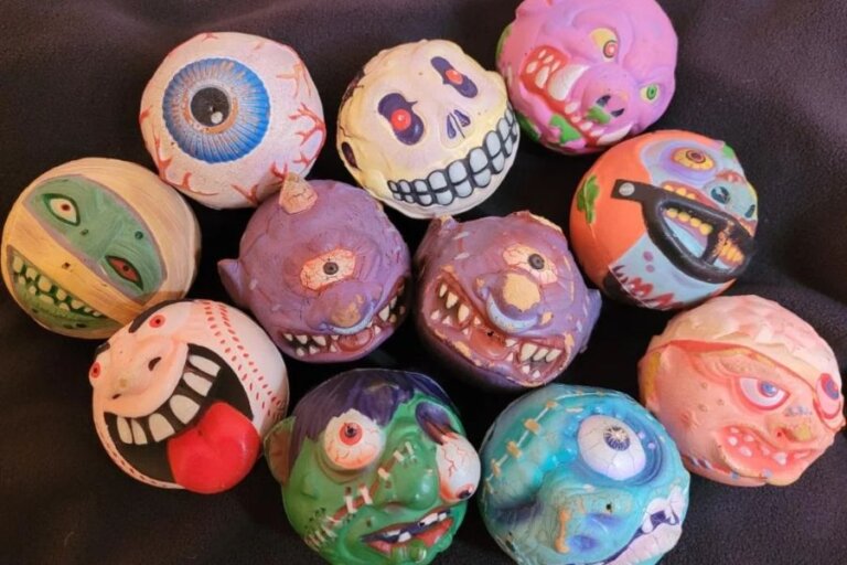 A collection of vintage Madballs foam toys is displayed on a dark fabric surface. Each ball features a grotesque, monster-like face with exaggerated details. The designs include a bloodshot eyeball, a grinning skull, and a baseball with a sinister expression. Other characters have sharp teeth, scars, stitches, and bulging eyes in vibrant colors.