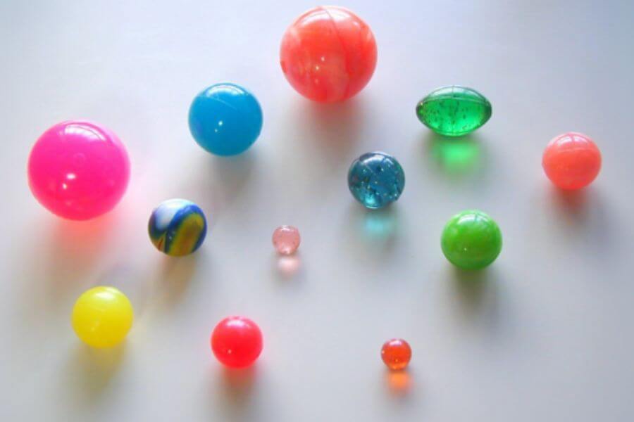 A collection of colorful bouncy balls is scattered on a smooth surface. The balls vary in size, with the largest being bright pink and orange. Some have a translucent, jelly-like appearance, while others are opaque and solid. A few feature swirls, glitter, or marbled patterns in shades of blue, green, and yellow. The glossy surfaces reflect light, emphasizing their rubbery texture. 