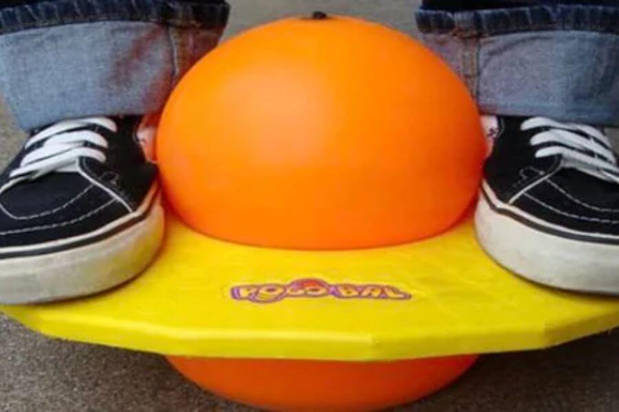 A classic Pogo Ball toy is being used on a concrete surface. The toy consists of an orange rubber ball squeezed between a yellow plastic platform. A person wearing black Vans sneakers and cuffed jeans is balancing on top. The Pogo Ball logo is visible on the platform, slightly worn from use. The bright colors contrast against the dull pavement.