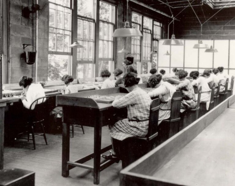 Black and white photo of a group of women working at long desks in a factory or workshop setting. They are seated in rows, focused on detailed tasks, with large windows providing natural light. Overhead lamps and a supervisor standing in the background suggest an early 20th-century industrial workspace.