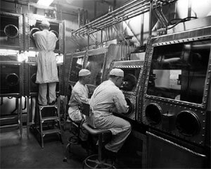 A black and white photograph of three workers in lab coats and caps handling materials inside sealed glove boxes in a laboratory. One worker is standing on a ladder, reaching into an upper containment unit, while the others sit at their respective stations. The specialized equipment suggests they are working with hazardous or radioactive substances in a controlled environment, likely in a mid-20th-century nuclear or radiochemistry facility.