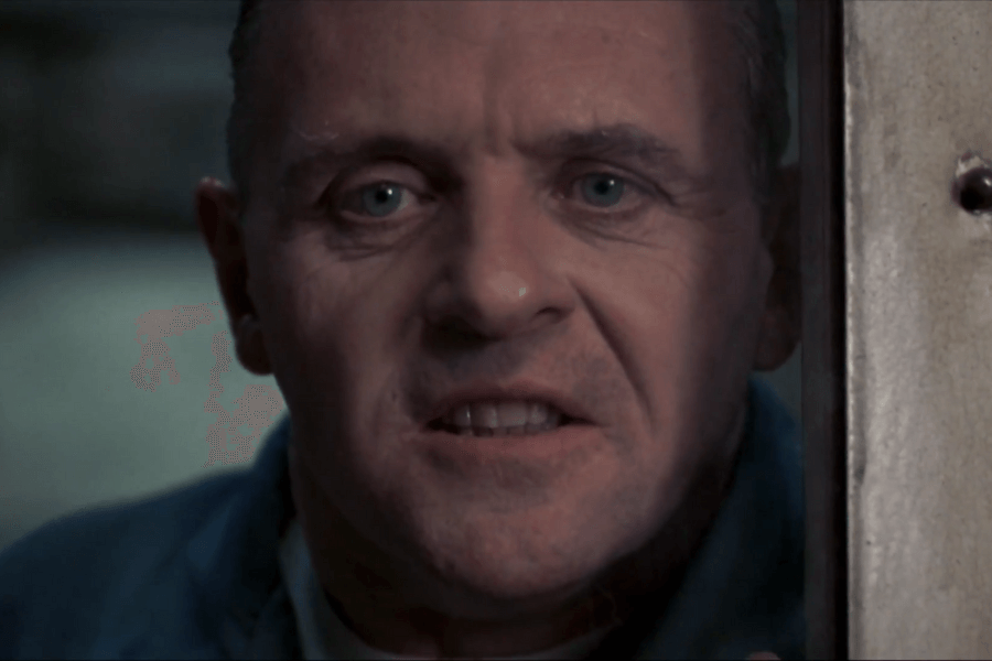 A man with piercing blue eyes and a chilling expression stares intently through a narrow gap in a metal door. His mouth is slightly open, revealing his teeth, as if he is speaking or about to make a sinister remark. The dim lighting and close-up framing enhance the unsettling intensity of the moment.