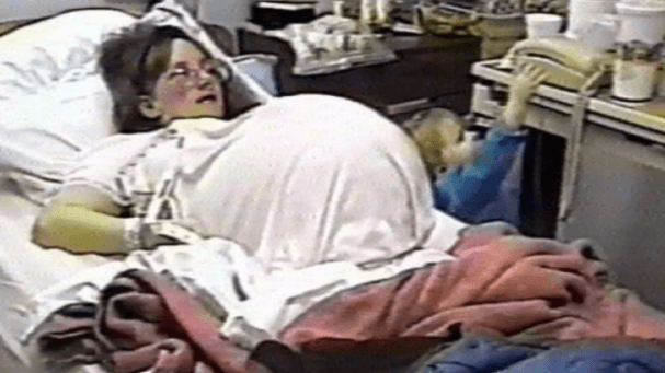 A pregnant woman with glasses lies in a hospital bed, her belly appearing extremely large under a white shirt. A small child in a blue outfit reaches for a telephone on a nearby table, while medical equipment and personal items are scattered around the room.