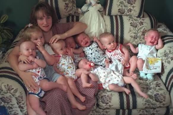 A mother sits on a floral-patterned couch, surrounded by seven babies dressed in colorful summer outfits. Some babies are crying, others are squirming, and one is holding a children's book, while the mother looks slightly overwhelmed but smiling.