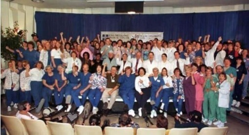 A large group of medical professionals, including doctors and nurses in scrubs, lab coats, and casual clothing, pose together in an auditorium with a blue curtain backdrop. Many are smiling, cheering, and raising their fists in celebration while an audience watches from the front rows.