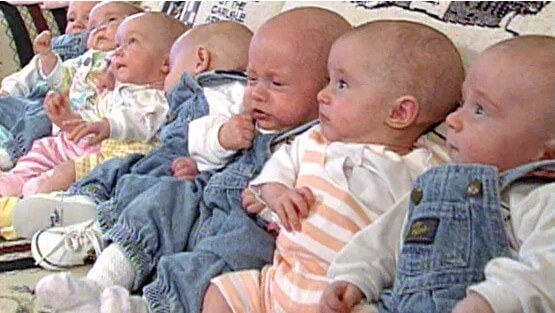 A row of seven babies dressed in various outfits, including denim overalls and striped onesies, sitting closely together. Some babies appear curious and alert, while others look sleepy or fussy.