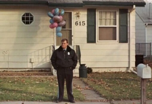 A uniformed police officer stands with hands in pockets outside a small beige house with green shutters, numbered "615." A cluster of pink and blue balloons is tied to the front porch railing, and a white mailbox sits near the sidewalk.