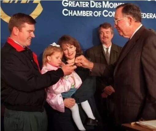 A man in a black and red jacket smiles while holding a young girl in a pink satin jacket, as an older gentleman in a suit hands something to her. A woman stands behind them, also smiling, with another man in a suit in the background, in front of a blue backdrop that reads "Greater Des Moines Chamber of Commerce Federation."