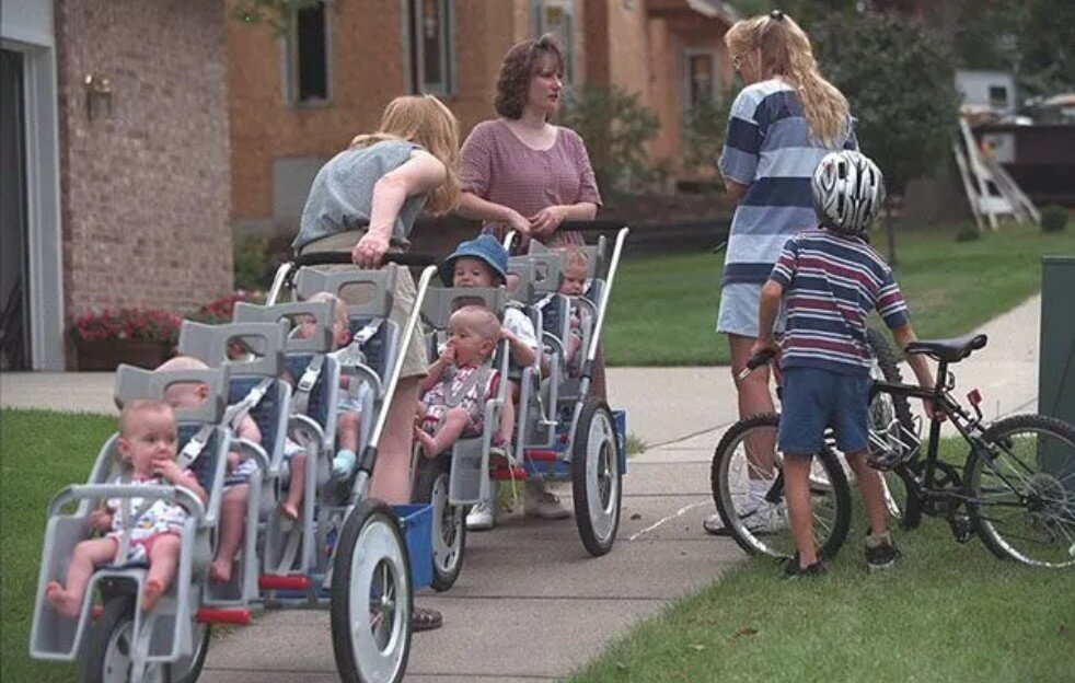 Two large multi-seat strollers filled with several babies are being pushed down a suburban sidewalk by two women, while another woman and a young boy with a bicycle stand nearby. The babies, dressed in colorful outfits, are secured in their seats, some looking around curiously while others suck on their fingers.