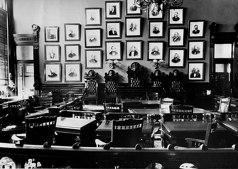 A black and white photograph of an old courtroom or council chamber with ornate wooden furniture and high-backed chairs arranged around desks. The walls are adorned with numerous framed portraits of distinguished figures, and a chandelier hangs from the ceiling, adding to the historic ambiance